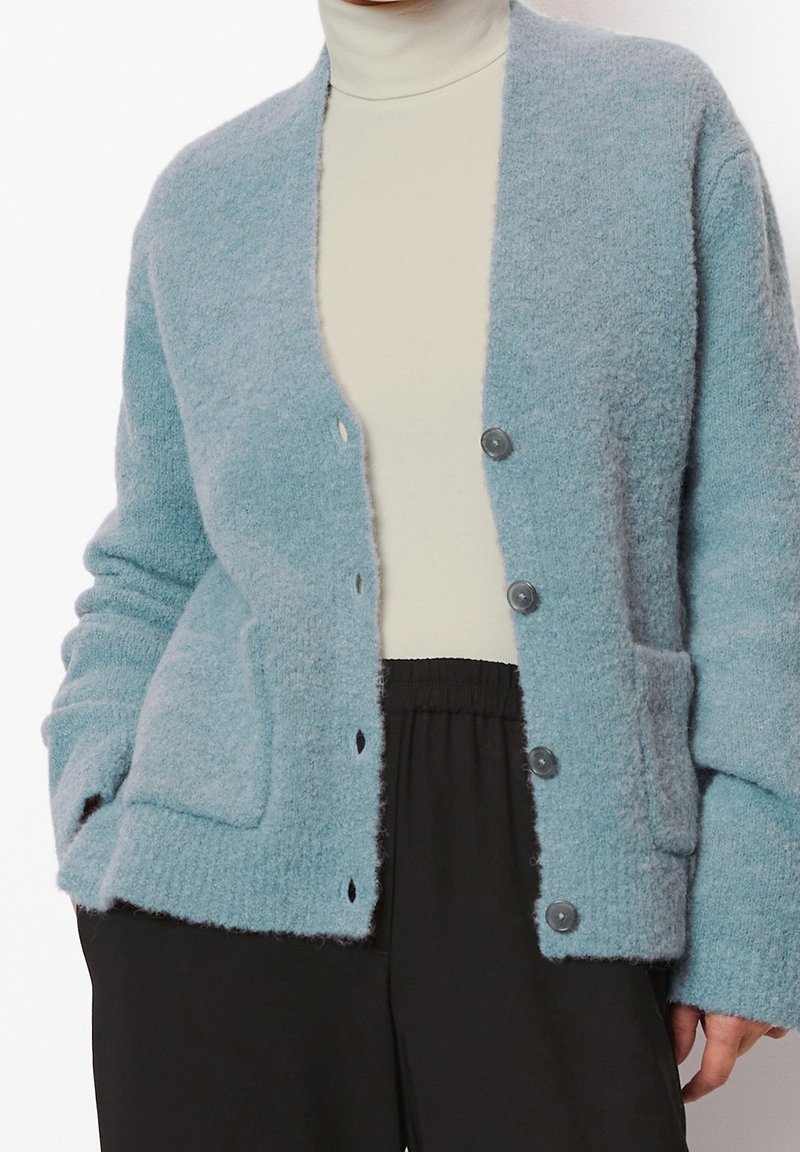 Cardigan bleu clair duveteux boutonné porté sur un col roulé crème et associé à un pantalon noir à taille élastique.