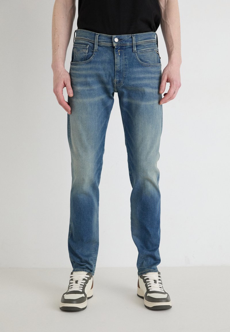 Modre jeans hlače z ozkim krojem, ki imajo svetlo bledenje, klasičen dizajn s petimi žepi in zgornji gumb. Oblečene z nevtralnimi teniskami.