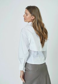 Camicia bianca con bottoni e maniche lunghe, dotata di un design posteriore a strati, abbinata a una gonna in pelle grigia aderente con tasche posteriori.