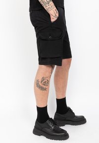 Zwarte cargoshorts met zijzakken, gemaakt van lichte stof. Dragen met zwarte sokken en schoenen, met een tatoeage zichtbaar op het been.
