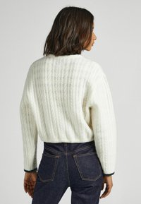 Maglione crema a treccia con taglio corto, colletto a coste e bordi scuri a contrasto. Abbinato a jeans di denim scuri a vita alta.