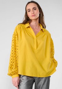 Camicia gialla con colletto a punta, chiusura a bottoni e maniche lunghe a pois. Il materiale sembra leggero, con una vestibilità rilassata e dettagli testurizzati.