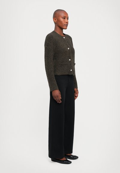 Short, dark green textured cardigan with front button closure and two pockets, paired with wide black trousers and black flats.