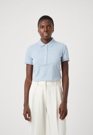 Hellblaues Polohemd mit Kragen, drei Knöpfen und kleinem Logo auf der Brust. Kombiniert mit cremefarbenen, hoch sitzenden Hosen.