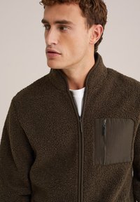 Chaqueta de forro marrón con cuello alto, cierre de cremallera y un bolsillo de tela contrastante en el lado izquierdo del pecho. Material texturizado y cálido.