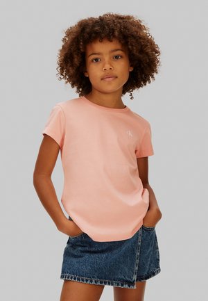 Child with curly hair wearing a light pink short-sleeve shirt and denim skirt, standing with hands in pockets against a plain background.