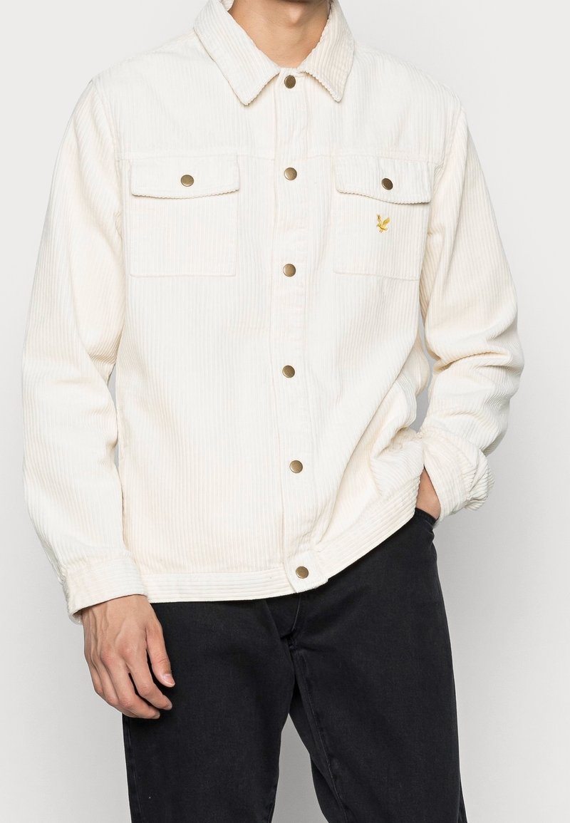 White corduroy jacket with two chest pockets, gold-tone buttons, and a small yellow emblem on the left side. Smooth texture, relaxed fit.
