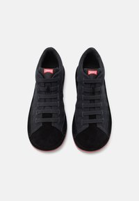 Schwarze Sneaker mit einem Wildleder-Obermaterial, strukturierten Details, sechs Ösen mit Schnürsenkeln und einem roten Logotag an der Zunge. Starre Gummisohle.