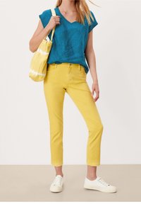Camiseta turquesa de cuello en v y mangas cortas, combinada con unos jeans amarillos cortos. Las zapatillas blancas y una bolsa amarilla de tie-dye completan el conjunto.