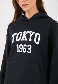 Zwarte katoenen hoodie met een voorste kangoeroezak en een witte grafische afbeelding met "TOKYO 1963" in vette letters.