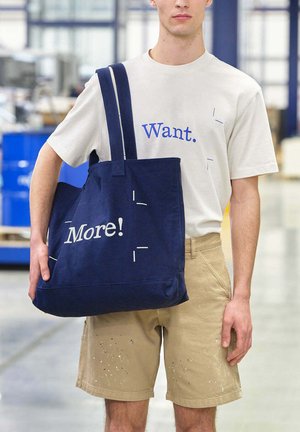 Giovane uomo con pantaloncini beige e t-shirt bianca con la scritta "Want." che porta una grande borsa blu navy con la scritta "More!" in un ambiente di magazzino industriale.