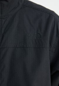 Svart, lett jakke med strukturert stoff. Har høy krage og et brodert logo på venstre bryst.