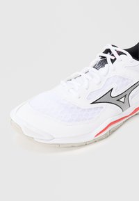 Άσπρο αθλητικό παπούτσι με επάνω μέρος από δίχτυ, μαύρες πινελιές, ασημένιες λεπτομέρειες και κόκκινη ρίγα στη σόλα. Διαθέτει παραδοσιακά κορδόνια και μαλακό σχεδιασμό.