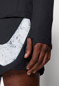 Svart langermet topp med ribbestruktur og tommelhull, kombinert med svarte shorts med et hvitt spraglete innslag i en uregelmessig form.