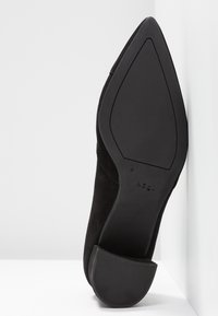 Chaussure à talon haut en daim noir, avec un bout pointu et une semelle lisse et plate. Le talon est de hauteur moyenne, sans motifs ni dessins visibles.