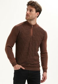 Brązowy sweter z zamkiem na szyi, z teksturowanym geometrycznym wzorem, prążkowanymi rękawami i dołem. Posiada gładką tkaninę i dopasowany krój.