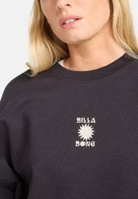 Felpa grigia scura con scollatura rotonda, polsini a costine e una grafica del sole ricamata bianca con il testo "BILLA BONG" sottostante.