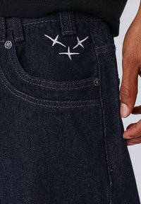 Jeans in denim scuro con ricami bianchi di tre uccelli vicino alla tasca, con vita classica e dettaglio di rivetti in metallo.