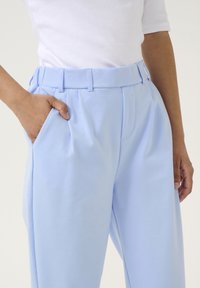 Pantaloni azzurri chiari con una texture liscia, una cintura alta, pieghe e tasche laterali, indossati con un top bianco aderente.