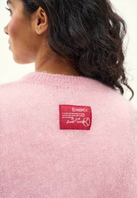Žena s tmavými kučeravými vlasmi, oblečená v huňatom ružovom svetri s červenou nášivkou obsahujúcou biely nápis „ROMANCE“ a krátkou potlačenou správou.