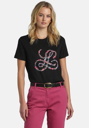 Kvinde iført en sort t-shirt med et flerfarvet blomster-svirveldesign, kombineret med lyserøde bukser med høj talje og et sort bælte med spænde i guld.