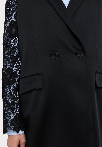Black blazer with dual buttons and large front pockets; features lace-embroidered sleeves with floral patterns; smooth fabric texture.