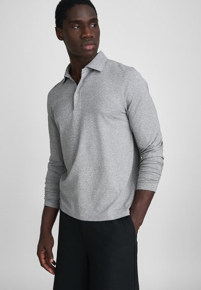 Polo gris à manches longues avec col, doté d'un plastron à boutons et de poignets côtelés. Tissu doux avec une coupe décontractée. Shorts noirs portés en dessous.