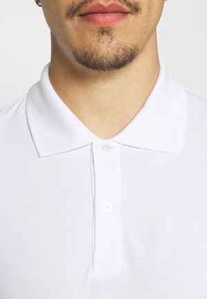 Unteres Gesicht und Hals einer Person mit kurzem Gesichtsbehaarung, die ein weißes Poloshirt mit Kragen und zwei zugeknöpften Knöpfen trägt.