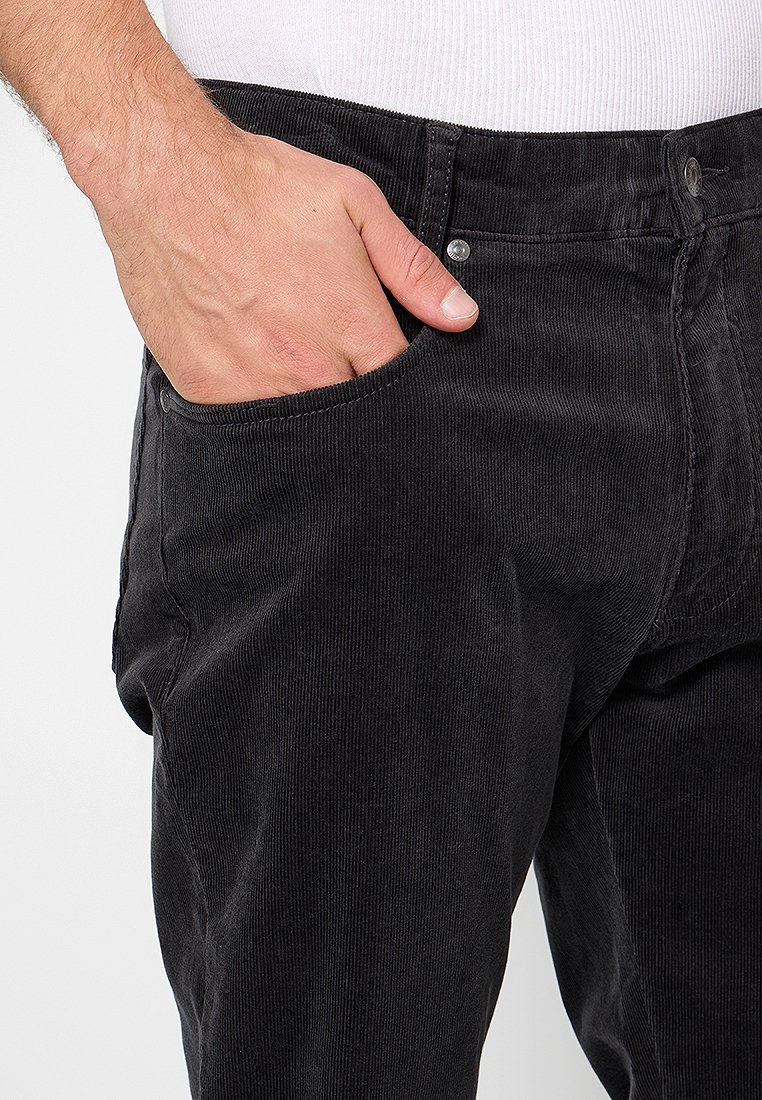 Männliche Hand, die in der Vordertasche von schwarzen Cordhosen ruht, die mit einem weißen Rippenhemd getragen werden.