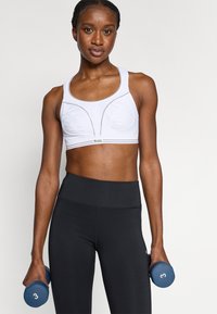 Vitt sport-bh med kurviga sömmar, kombinerad med högmidjade svarta leggings. Modellen håller blå hantlar märkta med "3".