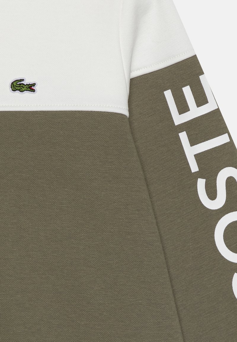 Sudadera en verde oliva y blanco con tejido texturizado, paneles contrastantes y gran texto en blanco "LACOSTE" en la manga. Presenta un logo bordado.