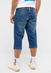Denim-Cargo-Shorts, knielang, leicht bis mittelblau gewaschen, mit zwei Gesäßtaschen und kontrastierenden Nähten. Tragen kombiniert mit weißen Sneakern und gemusterten Socken.