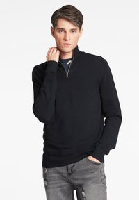 Schwarzer Pullover mit Reißverschlusskragen aus Strickstoff, mit langen Ärmeln und gerippten Bündchen. Trägt man über einem dunkelblauen Hemd mit lässiger Passform.