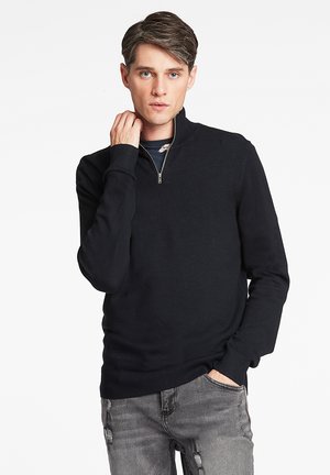 Jersey negro de cuello con cremallera, confeccionado en tejido de punto, con mangas largas y puños de canalé. Llevado sobre una camisa azul oscura de corte casual.