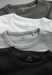 Drei Rundhals-T-Shirts gestapelt: schwarz, grau und weiß. Jedes hat eine gerippte Textur und ein Etikett mit der Aufschrift "The Set, Made in Bangladesh."