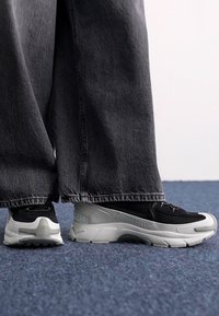 Black and gray sneakers with chunky soles, featuring a textured upper and unique design details on the heel and side. Worn with wide-leg gray jeans.