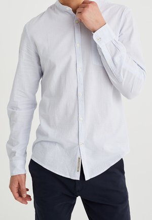 Mann trägt hellblau gestreiftes Hemd mit Stehkragen und navyblaue Hose, Hand nahe am Mund, vor einfarbigem Hintergrund.