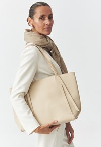 Beige nahkalaukku, jossa on jämäkkä muotoilu. Siinä on kaksi pitkää kantohihnaa ja hillitty logo. Mallilla on päälläsä vaalea huivi ja valkoinen bleiseri.