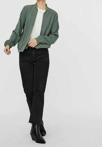 Chaqueta bomber verde claro con una textura suave, llevada sobre una camisa blanca, combinada con jeans negros de cintura alta y botas negras hasta el tobillo.