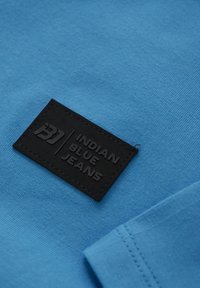 Blå bomullstyg med en svart gummimärke som har texten "INDIAN BLUE JEANS" och sömnadsdetaljer. Mjuk textur och enkel design.