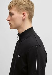 Svart zip-up jacka med hög krage, prydd med vita randdetaljer på ärmarna och en diskret logotyp på bröstet.