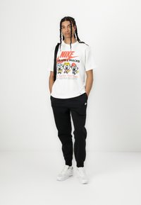 Hvit Nike t-skjorte med grafisk design som viser tegneseriefigurer, sammen med svarte Nike joggebukser og hvite sneakers.