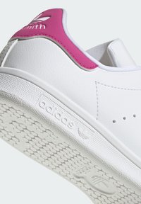 White sneaker with pink heel accent, smooth synthetic upper, textured white rubber sole, and perforations for breathability.