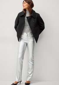Cappotto peacoat nero in lana con bottoni doppi, dolcevita grigia e pantaloni su misura argento lucido. Scarpe con tacco nere completano il look.