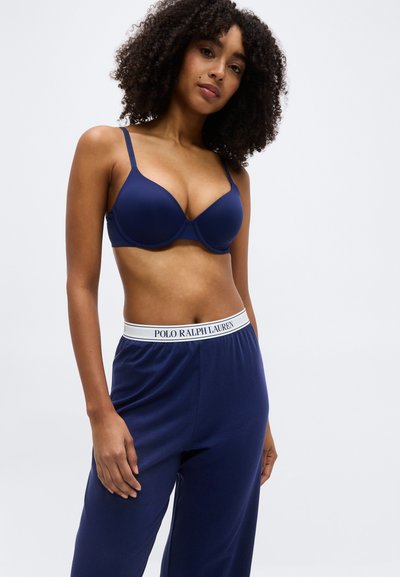 Γυναίκα με σγουρά μαλλιά που φοράει μπλε-σκούρο σουτιέν και μπλε-σκούρο παντελόνι Polo Ralph Lauren με λευκή μέση, σε απλό φόντο.