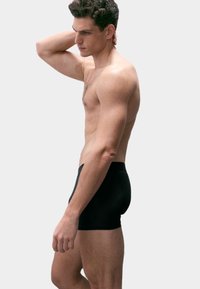 Černé boxerky vyrobené z pružného materiálu, s hladkou texturou a přiléhavým střihem, nošené mužským modelem ze stranového úhlu.
