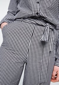 Mustavalkoinen gingham-ruudullinen asukokonaisuus, joka sisältää napillisen yläosan ja korkeavyötäröiset housut, joissa on solmittava vyö. Sileä kangaspinta.