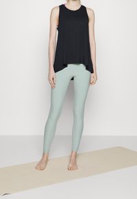 Osoba stojící naboso na béžovém jógovém koberci, oblečená v černém bezrukávovém topu a světlých zelených legínách na jednobarevném pozadí.