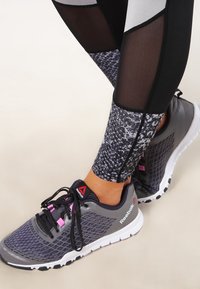 Scarpa sportiva Reebok in grigio con parte superiore in mesh nera e accenti rosa. Leggings neri e a fantasia con una sezione semitrasparente.