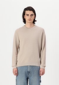 Maglione lungo beige a maniche lunghe con un design a maglia testurizzata, colletto rotondo e polsini. Abbinato a pantaloni in denim blu chiaro.